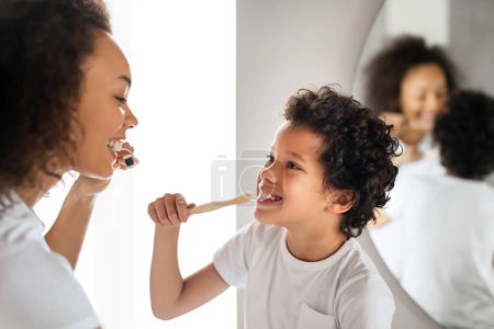 Téléchargez les photos : Un jeune garçon afro-américain, aux cheveux bouclés et au t-shirt blanc, sourit à une femme tout en apprenant à se brosser les dents. Elle démontre la bonne technique avec sa propre brosse à dents - en image libre de droit