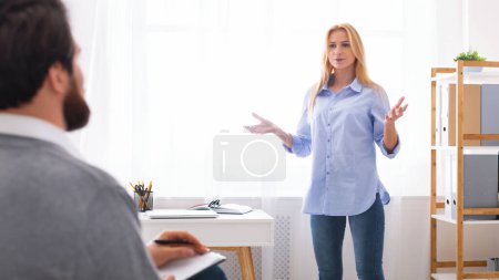 Emotional young blonde woman standing next to window at counselor office, expressing her feelings with man therapist