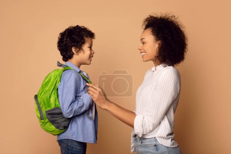Una alegre madre afroamericana arregla cuidadosamente las correas de la mochila de sus hijos mientras comparten un momento de afecto y conexión antes de que comience el día escolar.