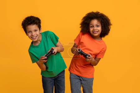 Téléchargez les photos : Deux jeunes enfants afro-américains, un garçon et une fille, se tiennent côte à côte avec de grands sourires, chacun tenant une manette de jeu vidéo, fond jaune - en image libre de droit