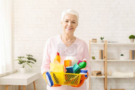 Die Seniorin steht und hält einen Korb mit verschiedenen Reinigungsmitteln wie Waschmittel, Scheuerbürsten, Schwämmen und Handschuhen in der Hand. Sie scheint bereit zu sein, das Haus zu putzen.