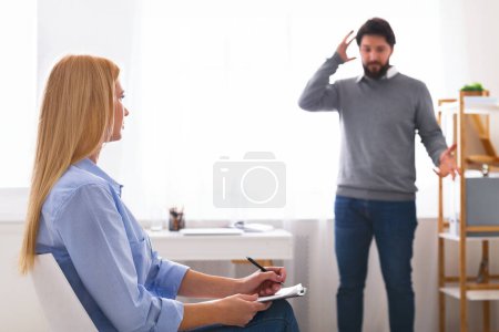 A man stands gesturing with his hand to his head in a sign of distress or making a point during a therapy session, as woman counselor, seated across from him