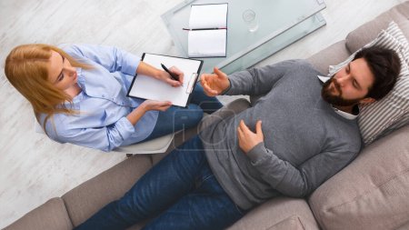 Photo for Top view of man client is reclining on a couch, discussing his thoughts with a woman therapist who is taking notes. The setting appears comfortable and private - Royalty Free Image