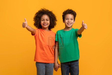 Photo for Two African American kids, one boy and one girl, are smiling and giving thumbs up gestures, bright yellow background. They appear cheerful and enthusiastic, showing approval in an expressive way. - Royalty Free Image