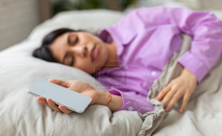 Téléchargez les photos : Une jeune femme du Moyen-Orient est capturée somnolant paisiblement dans un environnement de chambre confortable, vêtue d'un haut de pyjama violet décontracté, tenant un smartphone - en image libre de droit
