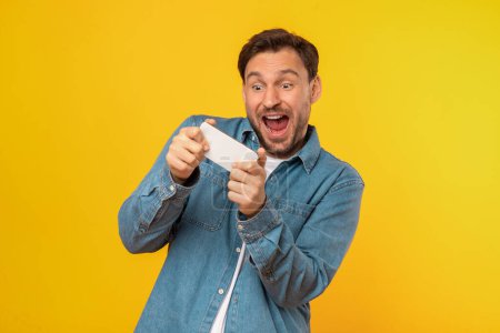 Foto de Un hombre adulto extático, luciendo una camisa de mezclilla casual, se para con una amplia sonrisa expresando euforia mientras juega en su teléfono inteligente, fondo amarillo - Imagen libre de derechos