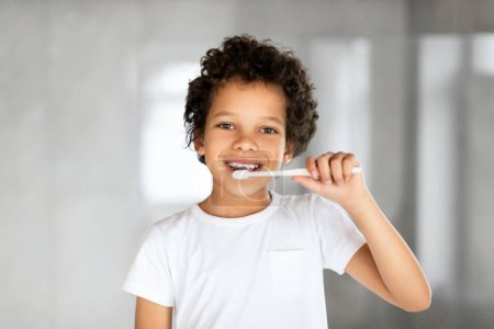 Téléchargez les photos : Afro-Américain jeune garçon aux cheveux courts se brosse les dents avec diligence avec une brosse à dents bleue devant un miroir. Il est debout dans une salle de bain, concentré sur sa routine d'hygiène buccodentaire. - en image libre de droit