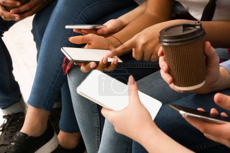 Photo for Cropped of diverse group of friends are seated closely together, each engrossed in their cell phones. They are holding the devices, fingers tapping and swiping on the screens - Royalty Free Image