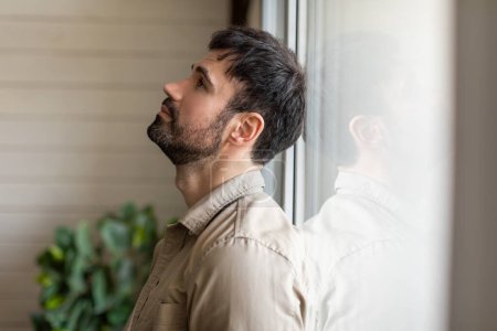 Photo for A man standing indoors beside a window, gazing outside at the scenery. The man appears contemplative as he observes the outdoor surroundings through the clear glass pane. - Royalty Free Image