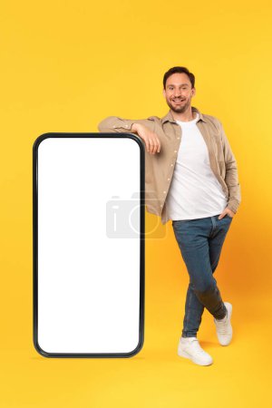 A smiling man in casual attire leans confidently against an oversized, blank white screen of a smartphone model, mockup copy space