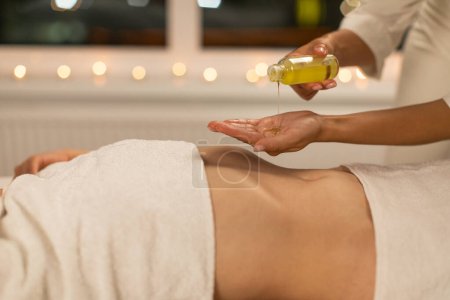 En un entorno de spa sereno con iluminación ambiental, se captura a una masajista profesional que se prepara para una sesión de masaje relajante vertiendo aceite de masaje en sus manos mientras el cliente espera.