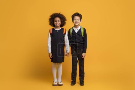 African American young girl and boy stand side by side, holding hands, ready for school. They are both dressed in smart uniforms, the girl in a pinafore and the boy in a vest and trousers