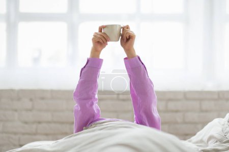 Mujer irreconocible sentada en una cama, sosteniendo una taza de café humeante en sus manos. Parece relajada, con los ojos cerrados, disfrutando de su rutina matutina..