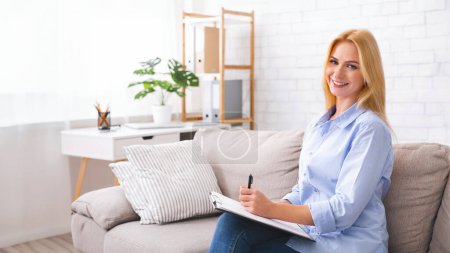 A woman with a bright smile, wearing a casual blue shirt, sits comfortably on a gray sofa holding a clipboard. She appears to be jotting down notes or completing a task