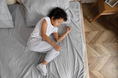Afroamerikaner mit lockigem Haar macht ein Nickerchen, zusammengerollt auf einem sauber gemachten grauen Bett mit weißer Bettwäsche, Draufsicht