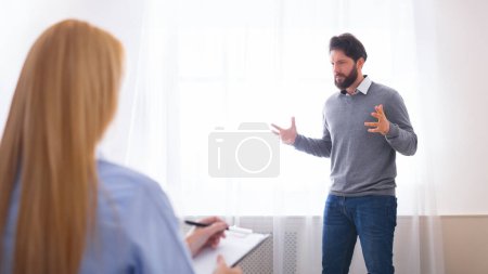 Hombre barbudo emocional de pie junto a la ventana en la oficina del consejero, haciendo gestos mientras habla, expresando sus sentimientos con la terapeuta mujer