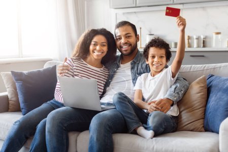 African American man and woman are seated on a couch, with a child holding a credit card in his hand. The family shopping online using laptop