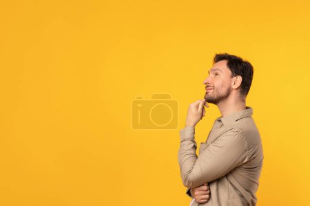 Photo for A pensive young man stands with his hand thoughtfully on his chin, looking upwards as if deep in contemplation against a bright yellow backdrop that highlights his reflective mood, copy space - Royalty Free Image