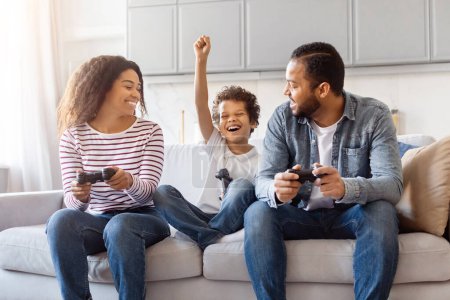 Eine fröhliche afroamerikanische Familie sitzt auf einem gemütlichen Sofa im Wohnzimmer und spielt. Das Kind wirft mit einem aufgeregten Blick eine Faust in die Luft, während es einen Controller hält