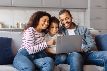 Photo for African American man, woman, and child are seated on a couch, all focused on a laptop screen in front of them. They appear engaged and concentrated on the digital content displayed. - Royalty Free Image