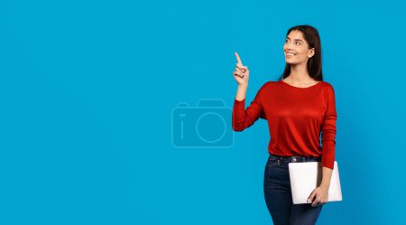 Téléchargez les photos : Une jeune femme joyeuse avec un sourire naturel se tient debout sur un fond bleu vif, regardant vers le haut et pointant vers le haut avec sa main droite, tenant un ordinateur portable, espace de copie - en image libre de droit