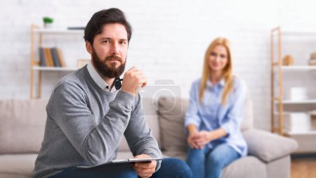 Photo for A bearded man therapist, holding a notebook, is seated attentively with a contemplative expression, while a lady client sits in the background on a couch - Royalty Free Image