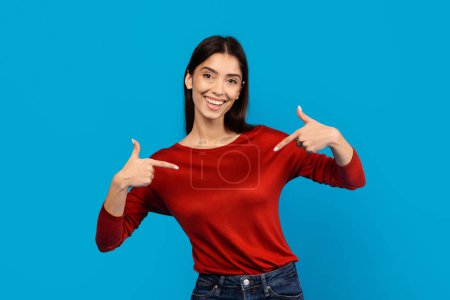A smiling young woman, wearing a casual red long-sleeved top, stands in front of a solid blue backdrop. With a look of self-assuredness, she points both thumbs toward herself