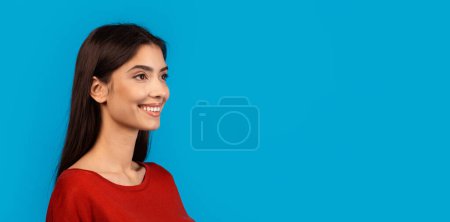 Photo for A young woman with long dark hair is seen in profile, wearing a bright red shirt against a vivid blue backdrop. She has a warm, engaging smile, looking at copy space - Royalty Free Image