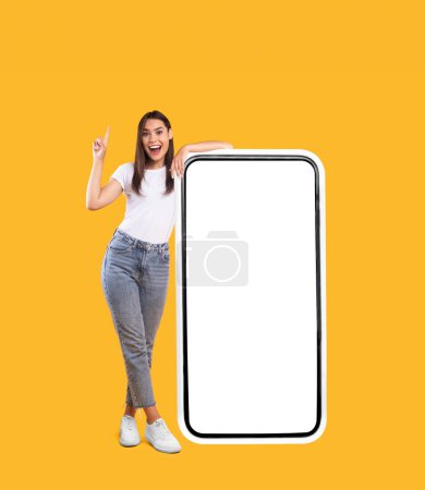 Wow. Happy Woman Leaning On Big Smartphone With Blank White Screen And Pointing Finger Up, Cheerful Lady Recommending New App Or Website, Standing On Yellow Background, Mock Up Image, Full Body Length