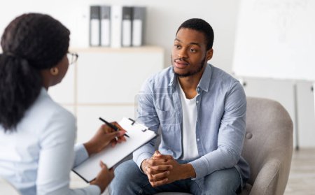 Foto de Unhappy young black man having session with professional psychologist at mental health clinic. Psychotherapist taking notes during conversation with depressed male patient - Imagen libre de derechos