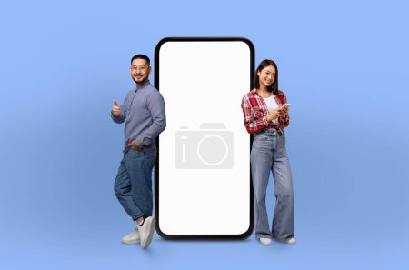 Téléchargez les photos : Couple asiatique debout à côté d'un téléphone de grande taille, regardant l'appareil avec curiosité. Le téléphone apparaît surdimensionné par rapport aux gens, créant un contraste visuel unique. - en image libre de droit