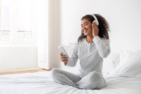 La mujer hispana está sentada en una cama, escuchando música a través de auriculares, sosteniendo el teléfono inteligente. Ella aparece enfocada y relajada mientras disfruta de su música en un entorno privado.