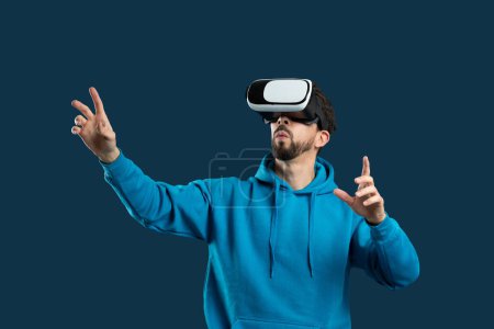 A man wearing a blue hoodie is immersed in a virtual reality experience through a headset. His hands are reaching out as if interacting with the virtual environment, blue background