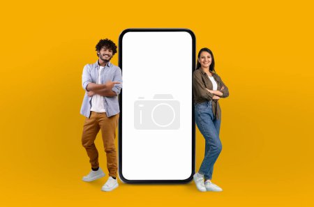 Un alegre joven hindú hombre y mujer de pie con confianza junto a un teléfono inteligente de pantalla grande y en blanco que domina el centro del marco, lo que sugiere un tema de tecnología