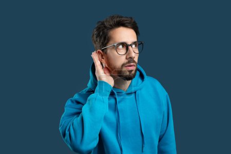 A man with a beard and glasses, dressed in a blue hoodie, stands against a solid color background. He appears focused as he cups his hand behind his ear, trying to hear something more clearly.