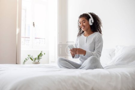 A young Hispanic woman sits cross-legged on her bed, wearing comfortable white pajamas and wireless headphones, engaged with her smartphone, possibly choosing a song or responding to a message