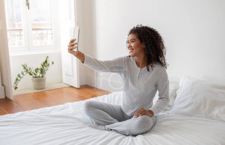 Foto de Mujer hispana sentada en la parte superior de una cama, sosteniendo un smartphone a lo largo de los brazos para capturar una selfie. Ella está enfocada en la pantalla, asegurando la toma perfecta mientras está sentada cómodamente en la cama. - Imagen libre de derechos