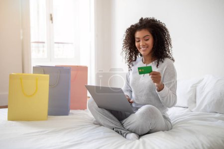 Hispanic woman is seated on a bed, holding a credit card in one hand and a laptop in the other. She appears focused on the laptop screen, possibly making an online purchase or managing finances.
