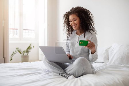 Hispanic woman is sitting on a bed, holding a credit card and a laptop in her hands. She appears to be engaged in online shopping or financial transactions.