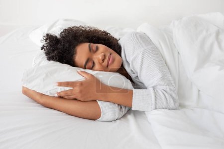 On voit une femme hispanique allongée au lit, avec un oreiller sous la tête. Elle semble confortable et détendue, peut-être reposante ou endormie, vue latérale