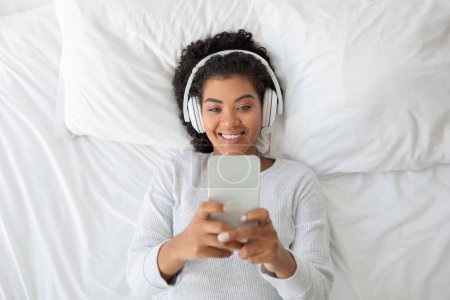 Hispanic woman lying down on a bed, actively engaged in listening to music through her smartphone. She appears relaxed and focused on her phone screen as she enjoys her music.