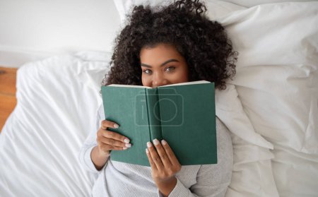 Hispanic woman is laying comfortably in bed, engrossed in a book she is reading. The room is softly lit, top view