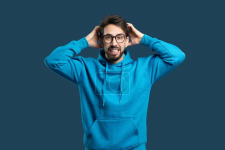 Ein Mann mit blauem Kapuzenpulli steht da, die Hände an die Ohren gedrückt, scheint in Not zu sein oder lautes Geräusch zu verspüren. Er wirkt angespannt und konzentriert auf die Geräusche, die er hört.