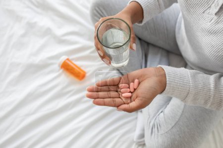 A woman holding a glass of water and a handful of pills in her hand. She appears to be taking medication according to the prescribed dosage, cropped