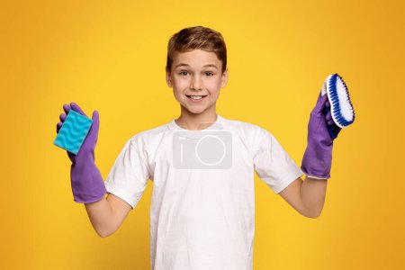 Téléchargez les photos : Mignon adolescent garçon en gants en caoutchouc tenant éponge et brosse, prêt pour le nettoyage de la maison, fond orange - en image libre de droit