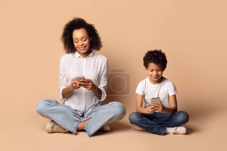 Afroamerikanerin und Kind sitzen auf dem Boden, beide intensiv auf einen Handybildschirm fokussiert. Sie scheinen in alles vertieft, was auf dem Gerät angezeigt wird.
