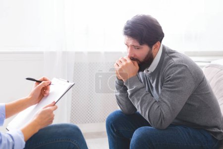 A distressed man is seen with his hand on his mouth as if in deep thought or concern, talking to a counselor who is taking notes. They are seated across from each other in a well-lit office space