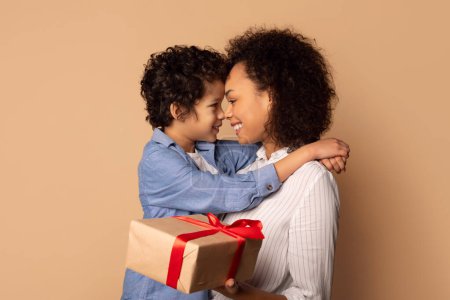 Téléchargez les photos : Mignon petit garçon afro-américain saluant sa mère avec la fête des mères, lui donnant boîte enveloppée et embrassant sa momie, fond studio beige - en image libre de droit