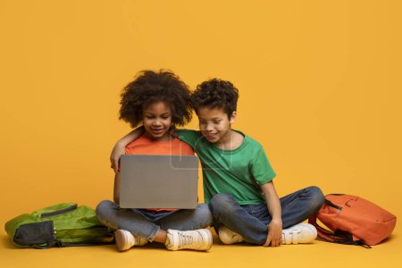 Téléchargez les photos : Deux jeunes enfants afro-américains assis sur le sol, engloutis dans un écran d'ordinateur portable. Ils sont concentrés, tapant et interagissant avec l'appareil sur fond jaune - en image libre de droit