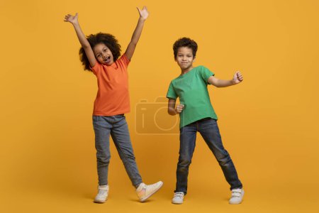 Two happy children, African American girl and boy, are dancing with raised arms, celebrating and showing excitement on yellow background